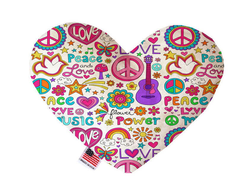 Heart shaped squeaker dog toy. White background with multicolored peace symbols, guitars, flowers, butterflies, doves, stars, and miscellaneous printed throughout. Made in USA label on bottom trim.
